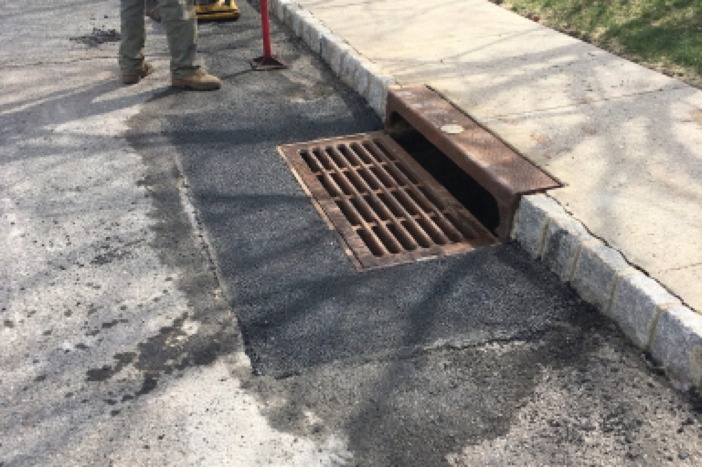 Beckage Sealcoating crew compacting a fresh asphalt patch around a reset storm drain grate in a NJ curb line
